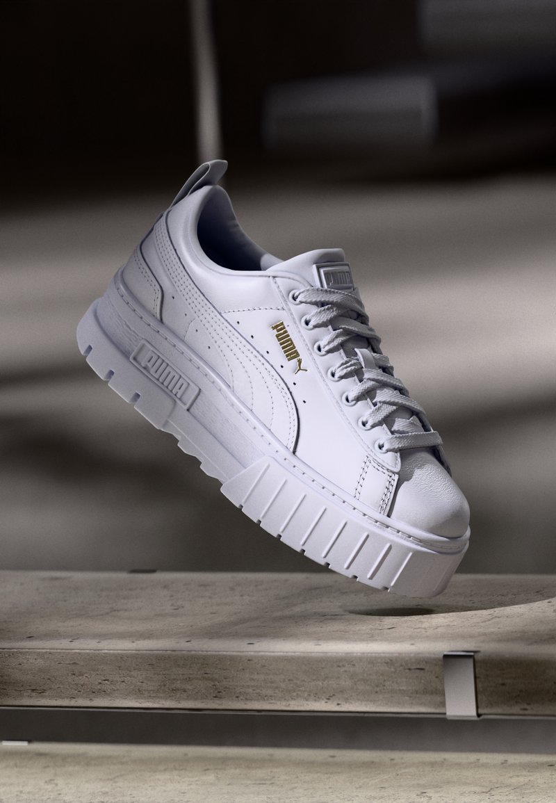 Baskets à plateforme blanches Puma avec une semelle épaisse, des lacets et un logo Puma doré, positionnées au-dessus d'une surface en béton avec un arrière-plan sombre flou.