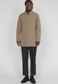 Trench de color marrón claro con cuello alto, cierre de botones y bolsillos laterales, combinado con pantalones y zapatos negros. Textura suave y diseño sencillo.