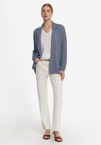 Stickad blå cardigan med sjalkrage, med ett strukturerat mönster och knappar, ihop med en vit V-ringad topp och lätta byxor.