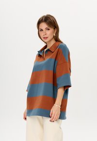 Oversized koszulka polo w brązowe i niebieskie poziome paski. Miękka bawełniana tkanina z kołnierzykiem i krótkimi rękawami. Luzny krój.