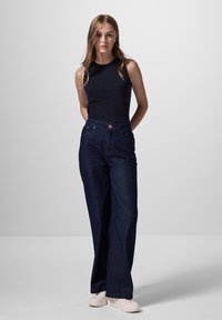 Jeans dritti blu scuro abbinati a un top nero fitted senza maniche. I jeans presentano un bottone frontale e una minima decorazione delle tasche.