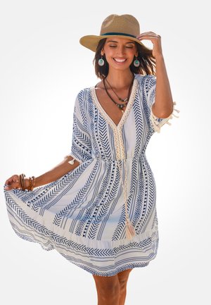 Biała sukienka z niebieskimi wzorami plemiennymi, dekoltem w serek, elastyczną talią i frędzlami. Stylizowana z słomkowym kapeluszem i warstwowymi naszyjnikami.