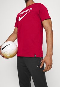 Punainen puuvillapaita, jossa lukee "LIVERPOOL FC" ja Nike-logo valkoisena. Harmaat puuvillahousut, joissa on pieni valkoinen Nike-logo. Henkilö pitää valkoista ja kultaa jalkapalloa.