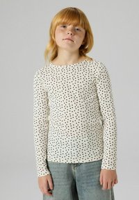Długorękawowa koszulka w kolorze kremowym z czarnymi groszkami, wykonana z prążkowanej dzianiny. Okrągły dekolt i dopasowany krój, zestawiona z jasnoniebieskimi dżinsami.