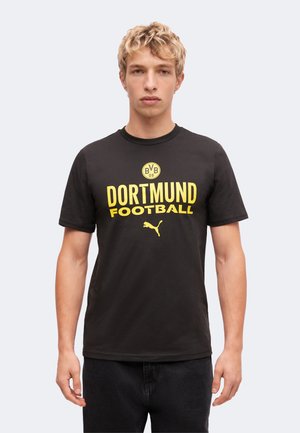 Ung mand med blondt hår iført sort Dortmund fodbold t-shirt med gult logo og Puma-symbol, står mod en ensfarvet baggrund.