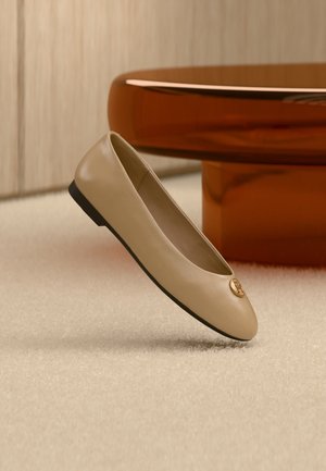 Ballerina beige in pelle con suola nera ed emblema dorato sulla punta, posizionata su tappeto crema accanto a un mobile di colore ambra arrotondato.