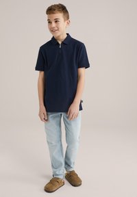 Tricou polo navy cu guler cu fermoar, asortat cu blugi albaștri deschiși și pantofi brown slip-on. Design simplu, material ușor.