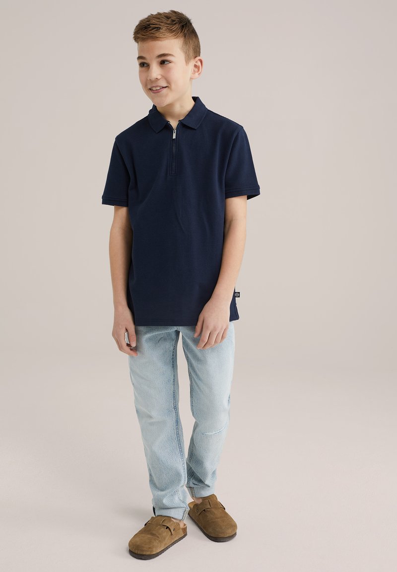Tricou polo navy cu guler cu fermoar, asortat cu blugi albaștri deschiși și pantofi brown slip-on. Design simplu, material ușor.