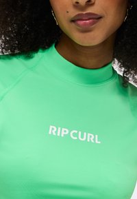 Femme aux cheveux bouclés portant un haut rash guard Rip Curl vert vif, montrant les lèvres, le menton, ainsi qu'une partie de son cou et de ses épaules.