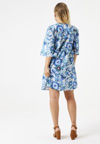 Kurzes Kleid mit kurzen Ärmeln in einem blauen und grünen abstrakten Muster, mit einer taillierten Taille und einem weit schwingenden Rock. Braune klobige Absätze runden den Look ab.