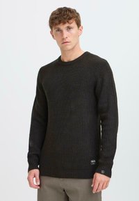 Dunkel gerippter Strickpullover mit langen Ärmeln und Rundhalsausschnitt. Verfügt über eine strukturierte Oberfläche und ein kleines Logoschild am unteren Saum.