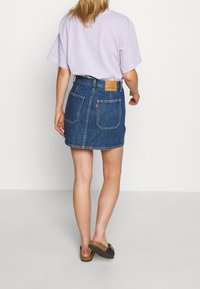 Falda de mezclilla azul, diseño ajustado, por encima de la rodilla, con dos bolsillos traseros y una etiqueta de cuero en la cinturilla. Lleva una camiseta lila.