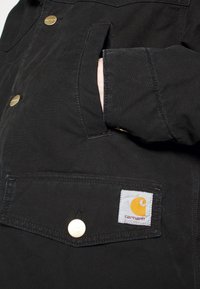 Dłoń częściowo włożona do czarnej kieszeni kurtki z widocznymi złotymi guzikami oraz białą naszywką z logo Carhartt na dolnej klapce kieszeni.