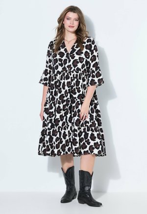 Kobieta w luźnej sukience w panterkę do kolan z rękawami 3/4 i czarnych kowbojskich butach, stojąca przy gładkiej ścianie.