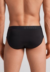 Sort brief med et glat, strækbart stof. Har en bred talje med præget logo. Formet til en tætsiddende støtte.
