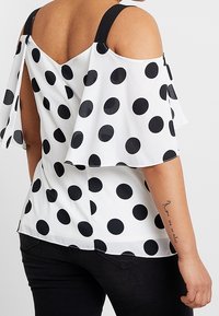 Blusa bianca a spalle scoperte con grandi pois neri, spalline nere e maniche a campana, indossata con pantaloni neri, che mostra un tatuaggio sul braccio.