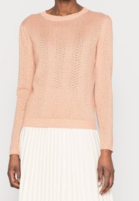 Pull en maille rose clair à manches longues, texturé avec des motifs ajourés, doté d'un col côtelé et de poignets festonnés.