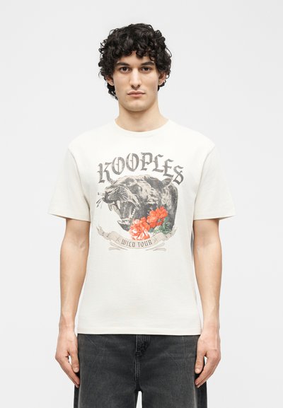 Άνδρας με σγουρά μαλλιά φοράει λευκό μπλουζάκι με εκτυπωμένη μια βρυχώμενη μαύρη πάνθηρα, κόκκινα λουλούδια και το κείμενο "KOOPLES WILD TOUR".