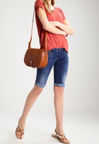 Mujer con pantalones cortos de mezclilla arremangados, camiseta coral de manga corta, bolso marrón cruzado y sandalias marrones, de pie con los brazos cruzados.