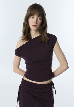 Jeune femme aux cheveux châtains mi-longs porte un haut asymétrique violet foncé et une jupe froncée assortie sur un fond uni.