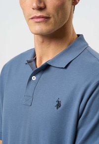 Modré polokošeľa s jemnou textúrou, vybavená troma gombíkmi na zapínanie a vyšitým čiernym logom na ľavej strane hrudníka.