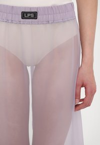 Světlé levandulové průhledné kalhoty se strečovým pasem, s viditelnou černou etiketou. Materiál je lehký, poloprůhledný a hladký.