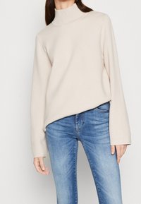 Pull col roulé beige clair à manches longues, texture douce et coupe décontractée, associé à un jean en denim bleu.