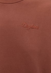 Gros plan sur un tissu marron avec le logo "Drykorn" brodé en fil assorti près de l'encolure.