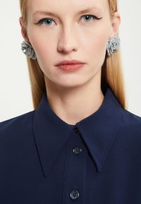 Námořnická košile na knoflíky s špičatým límcem, vyrobená z texturované látky. Ušnice s jemnými stříbrnými korálky tvoří dekorační akcent.