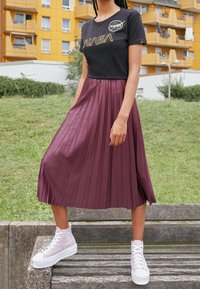 Svart NASA-logotyp t-shirt, plisserad bordeaux midi-kjol, och ljusgrå högskor med vita sulor, placerad på en träbänk utomhus.