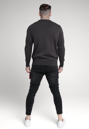 Dunkelgrauer Pullover mit gerippter Textur, eng anliegenden Ärmeln und Rundhalsausschnitt. Kombiniert mit schwarzen Skinny-Jeans und weißen Sneakern.