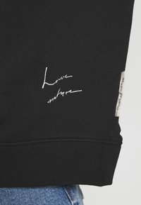 Sudadera negra con texto bordado en blanco "Ama la naturaleza", puños acanalados y una etiqueta de tela con "Marc O'Polo" en el costado.