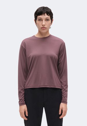 Personne aux cheveux courts et foncés portant un haut mauve à manches longues et un pantalon noir, debout devant un fond clair uni, regardant vers l'avant.