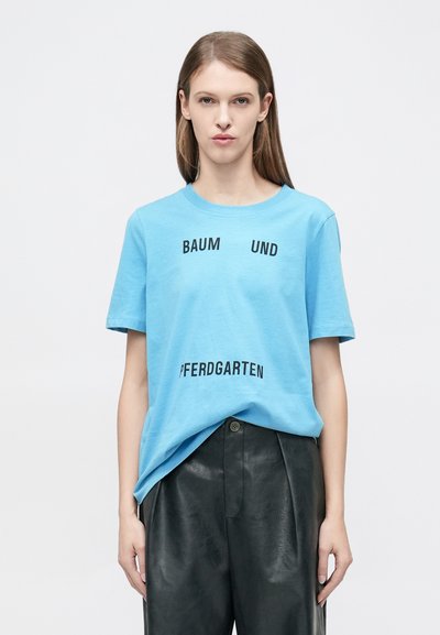Ανοιχτό μπλε βαμβακερό T-shirt με μαύρο κείμενο που γράφει: "BAUM UND" και "PFERDGARTEN." Κοντά μανίκια, στρογγυλή λαιμόκοψη, άνετη εφαρμογή.