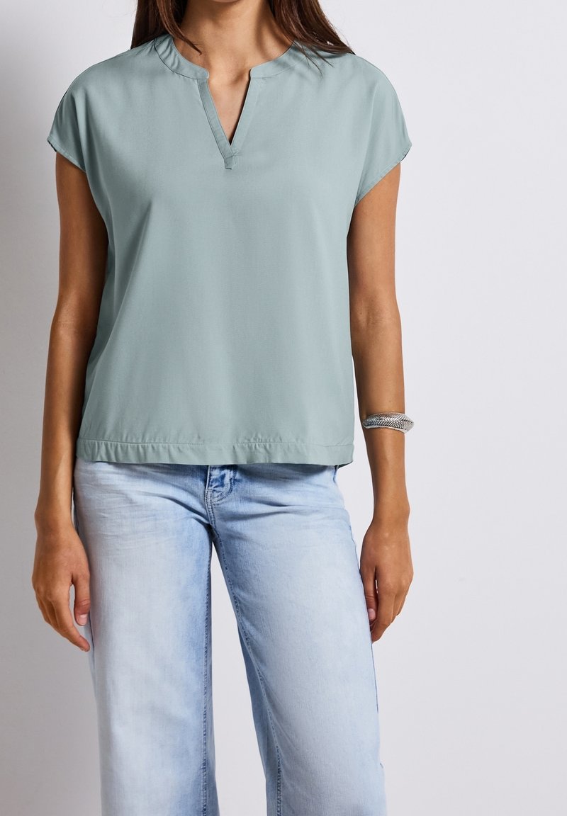Femme portant un chemisier bleu clair à manches courtes avec une entaille en V et un jean délavé, bracelet argenté au poignet droit, debout devant un fond gris.