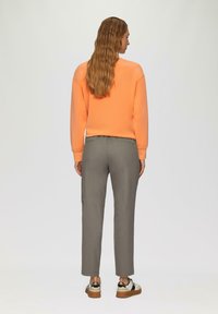Oranje sweatshirt en grijze slim-fit broek. Schoenen hebben een contrasterend ontwerp. Haar is in losse golven gestyled. Eenvoudige achtergrond.