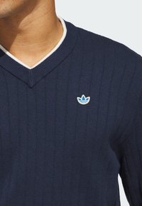 Navy ribbestrikket genser med V-hals, med hvite detaljer rundt kragen og en liten blå og hvit brodert logo på brystet.
