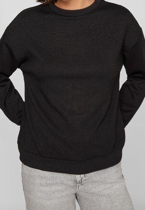 Personne portant un sweat-shirt noir uni à manches longues avec des poignets côtelés et un jean gris clair sur un fond uni.