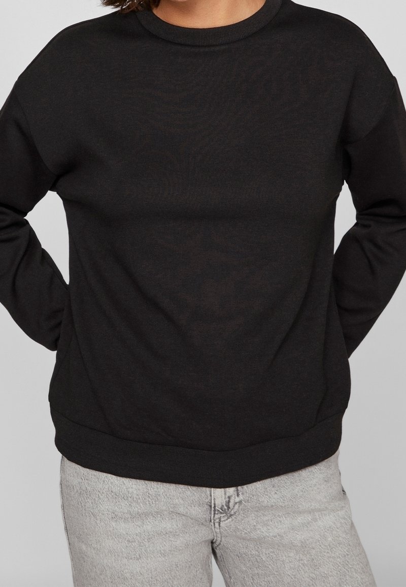 Personne portant un sweat-shirt noir uni à manches longues avec des poignets côtelés et un jean gris clair sur un fond uni.