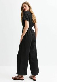 Combinaison noire à manches courtes, jambes larges et taille ceinturée, fabriquée en tissu texturé. Complétée par des sandales plates marron.