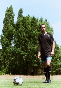 Czarna sportowa koszulka z szarymi akcentami, niebieskie szorty i czarne skarpety. Stoi na trawie obok wielokolorowej piłki do soccera i drzew.