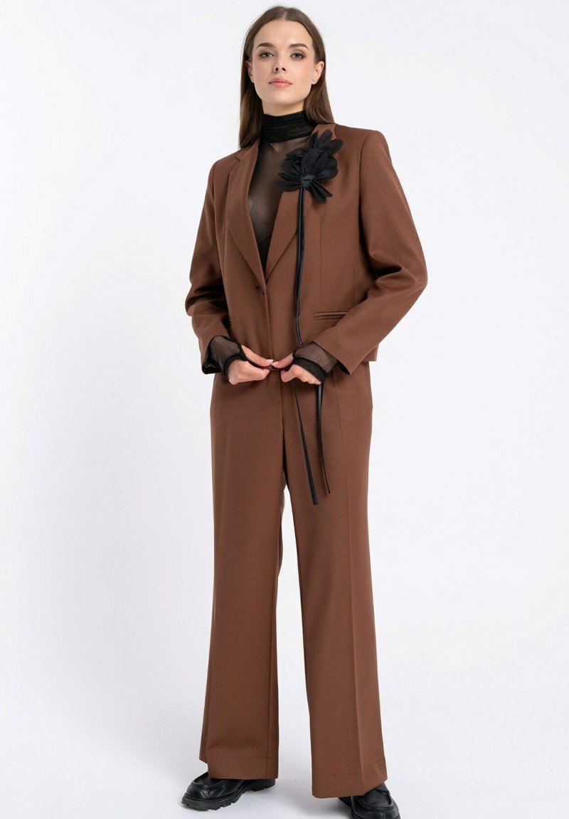 Brązowy garnitur szyty na miarę z prostymi spodniami, blazerem na jeden guzik i akcentem w postaci czarnej, jedwabnej kwiatu. Przezroczysta czarna bluzka pod spodem.