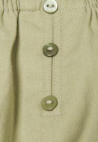 Detail béžové tkaniny s třemi svisle uspořádanými zelenými knoflíky, které jsou přišity béžovou nití.