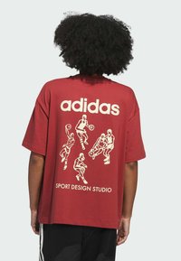 Červené oversized tričko s bílým logem "adidas" a grafikou basketbalisty na zádech. Krátké rukávy, bavlněná tkanina, volný střih.