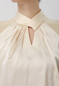 Kremfarvet satinbluse med høj krave, knude og plisseringer, med en nøglehulsløsning foran under halsen.
