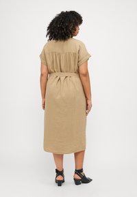 Khakifarbendes Leinen-Midikleid mit kurzen umgeschlagenen Ärmeln, tailliertem Gürtel und gerafftem Rückendetail. Kombiniert mit schwarzen Absatzsandalen mit Knöchelriemen.