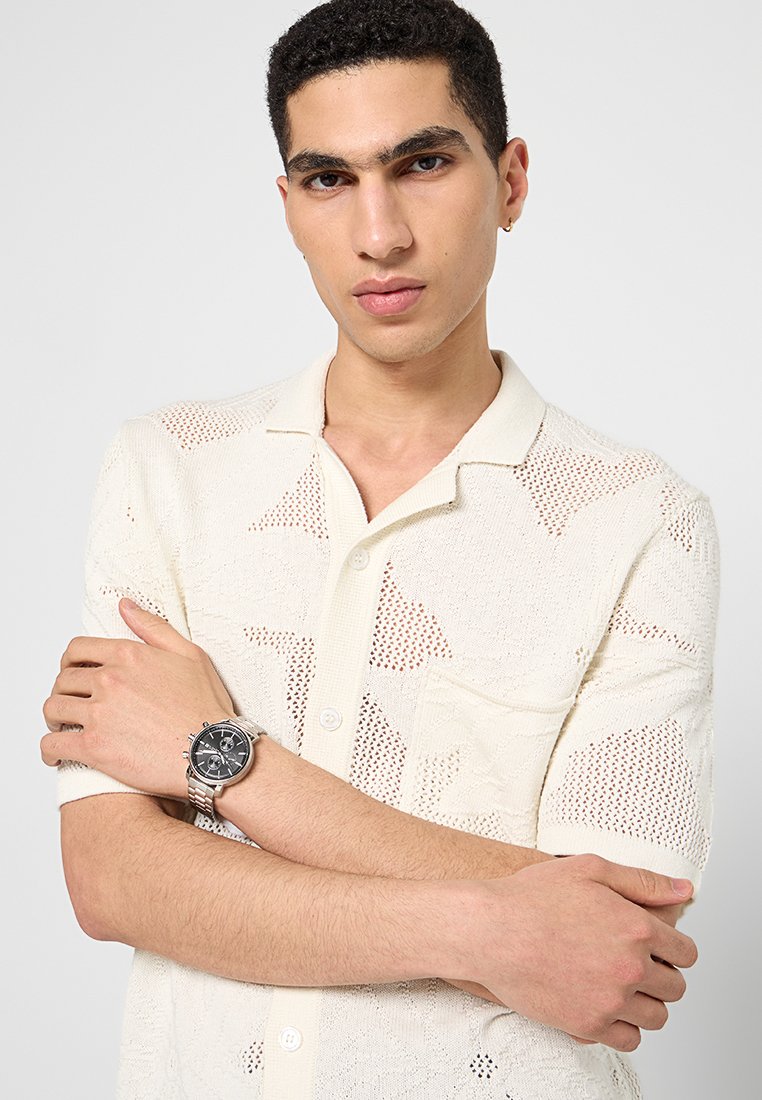 Gestricktes Kurzarmhemd in Creme mit einem strukturierten Muster, ausgestattet mit einem Kragen und Knöpfen. Mann mit einer silbernen Uhr am Handgelenk.