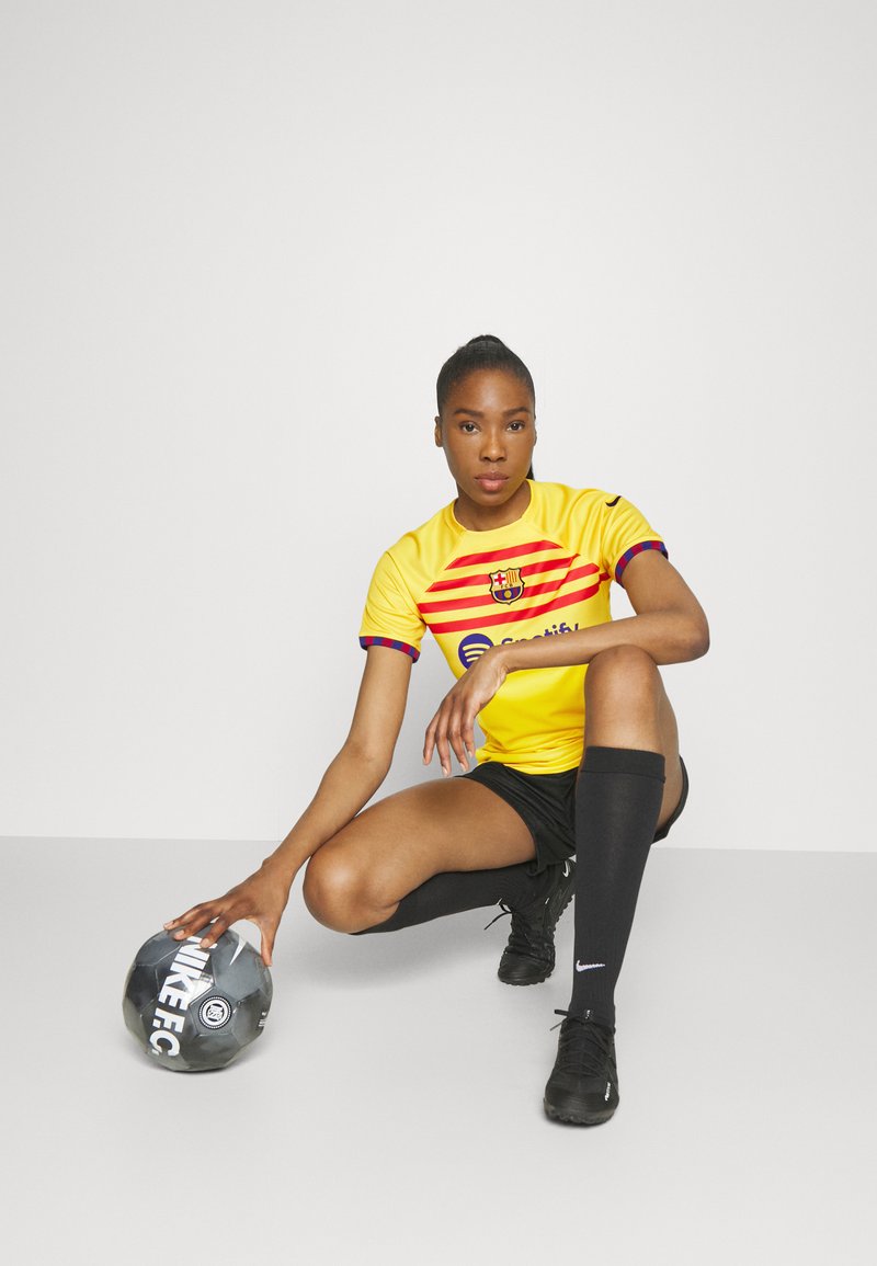 Camiseta de fútbol en amarillo brillante con franjas rojas y azules, pantalones cortos negros, medias largas negras y un balón negro marcado como "NIKEFC".