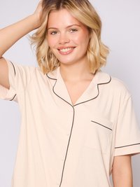 Beige kortærmet pyjamashirt med sort piping, knaplukning foran og en enkelt brystlomme. Glat tekstur, afslappet pasform.