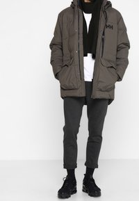 Giacca imbottita verde scuro con cappuccio, chiusura con zip e dettaglio del logo. Indossata sopra una camicia bianca, pantaloni grigi e calzature nere.